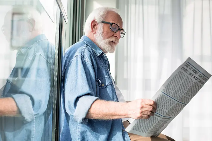 Älterer Mann mit weißem Bart und Brille lehnt entspannt an einer Fensterfront und liest konzentriert eine Zeitung.